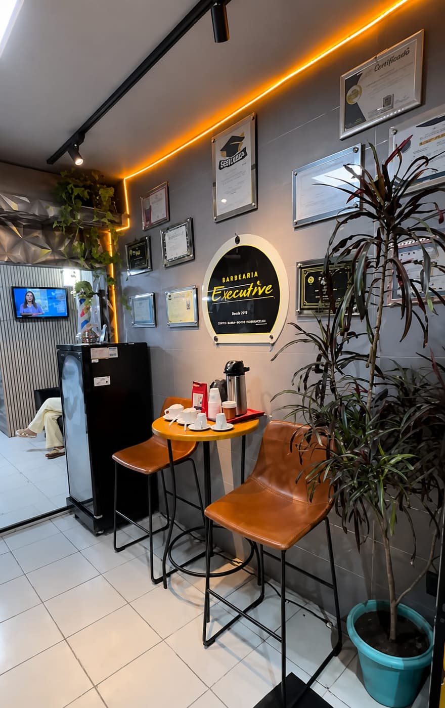 Espaço de espera da Barbearia Executive: mesa de café, TV e a parede de certificados de 30 anos de profissão